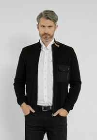 Czarna rozpinana kardigan z teksturowanym splotem, z kieszenią na piersi, brązowym akcentem przy kołnierzyku, noszony na białej koszuli z guzikami.