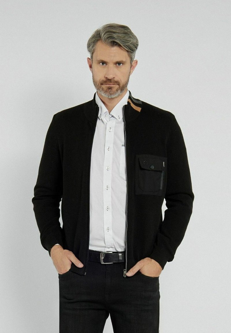 Czarna rozpinana kardigan z teksturowanym splotem, z kieszenią na piersi, brązowym akcentem przy kołnierzyku, noszony na białej koszuli z guzikami.