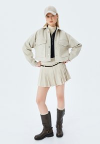 Hellgraue cropped Jacke mit zwei vorderen Taschen, beigefarbener Faltenrock, hohe braune Stiefel mit Schnallen und eine passende Mütze. Glatte Texturen überall.