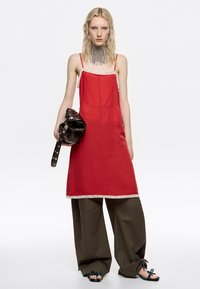 Młoda kobieta ubrana w czerwoną sukienkę na ramiączkach na szerokich oliwkowych spodniach, trzymająca złożoną ciemną skórzaną kurtkę, z dużym srebrnym naszyjnikiem łańcuchowym i sandałami.