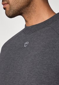 Σκούρα γκρι φούτερ με υφή υφάσματος, ριμπ στρογγυλή λαιμόκοψη και μικρό λευκό κεντητό λογότυπο στο στήθος.