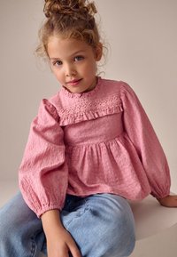 Roze blouse met ruches en een getextureerde stof, lange opgeblazen mouwen en een kanten rand bij de halslijn, gecombineerd met lichtblauwe spijkerbroek.