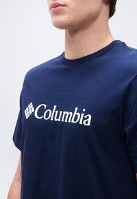 Gros plan d'une personne portant un t-shirt bleu marine avec le logo blanc Columbia et un emblème en forme de losange sur la poitrine.