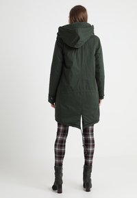 Žena v tmavě zelené parce s kapucí, černobílých a červenobílých kostkovaných legínách a tmavě šedých podpatkových kotníkových botách, otočená zády.