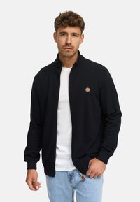 Hombre vistiendo una chaqueta negra con cremallera sobre una camiseta blanca y jeans de mezclilla azul claro, de pie frente a un fondo claro y liso.
