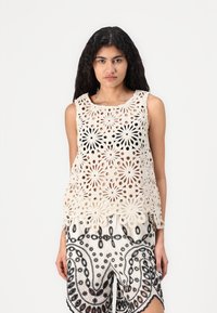 Top senza maniche all'uncinetto color crema con motivo floreale e design a traforo; indossato sopra pantaloni corti a motivi in bianco e nero.