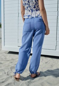 Pantalon bleu clair, ample, en tissu texturé, avec des poignets froncés aux chevilles et des poches arrière. Porté avec un haut à motif floral.