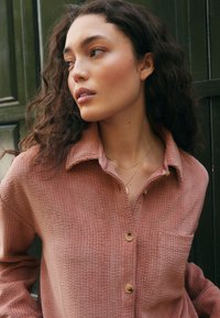 Camicia a bottoni rosa con texture, colletto, taschino singolo sul petto e bottoni in legno. Tessuto liscio e morbido con sottili righe a coste.