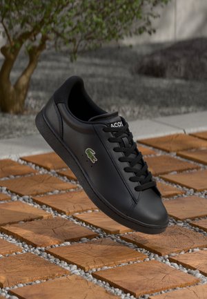 Chaussure de sport en cuir noir avec une finition lisse, dotée de lacets, d'un bout arrondi et d'un petit logo vert sur le côté, flottant au-dessus d'un sol carrelé.