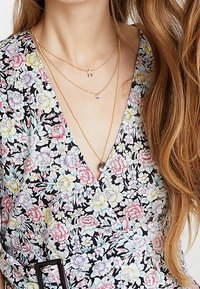 Collares de oro en capas que presentan un pequeño encanto y un colgante de cristal, combinados con un vestido negro de patrón floral con escote en V.