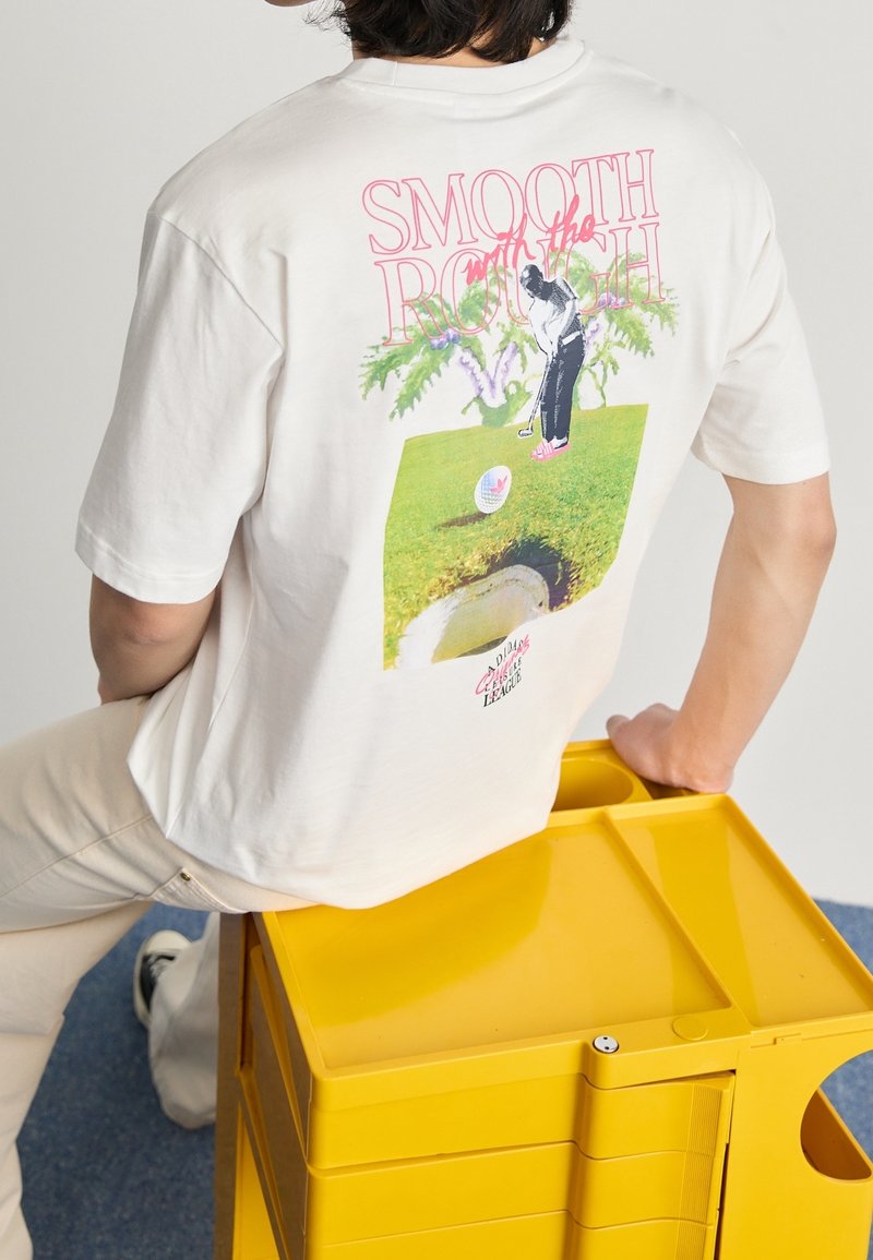 Valkoinen puuvillapaitapaito, jossa on graafinen kuvasto selässä: golfari, golfpallo ja trooppisia elementtejä. Lyhyet hihat, rento istuvuus.