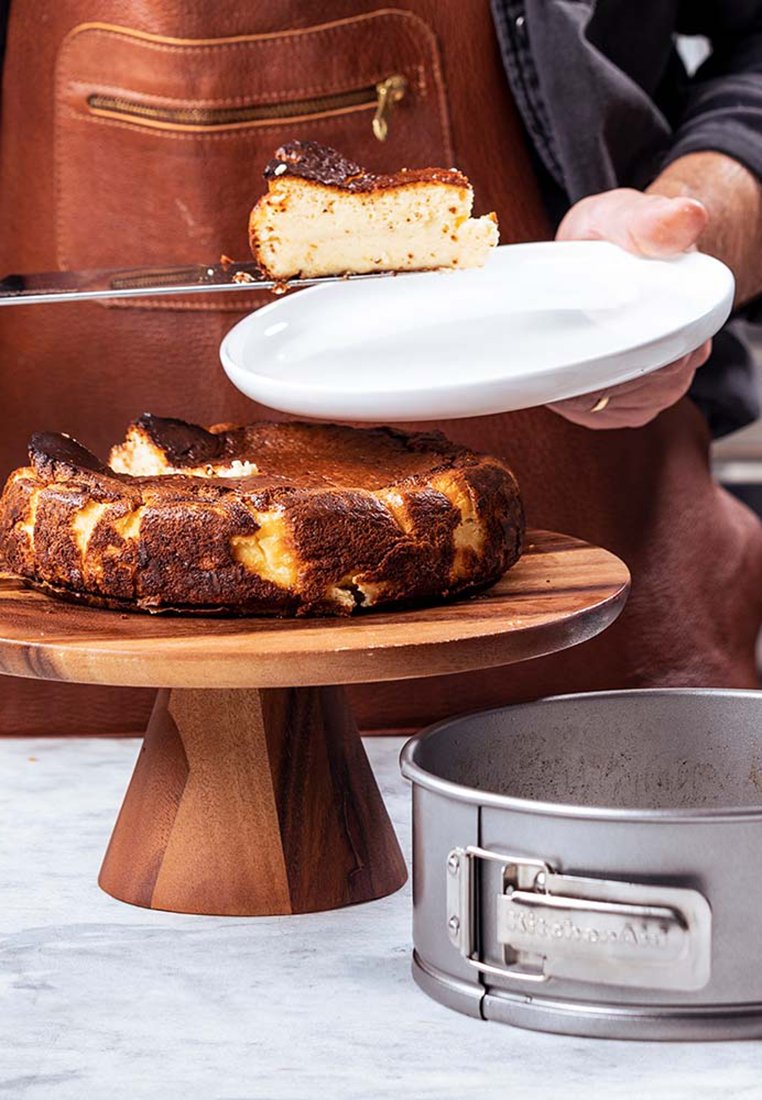 Ronde, donkerbruine cheesecake op een houten voetstuk, met een kruimelige bovenkant. Een stuk wordt geserveerd op een wit bord, naast een springvorm.