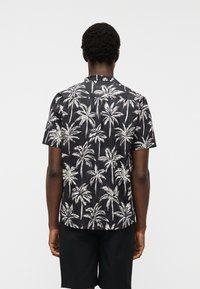 Άνδρας που φοράει μαύρο κοντομάνικο πουκάμισο με λευκό μοτίβο φοίνικα και μαύρο σορτς, στέκεται με την πλάτη προς την κάμερα μπροστά σε απλό φόντο.