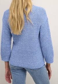 Light blue knitted sweater with three-quarter sleeves, ribbed hem, and textured pattern. Paired with light denim jeans.