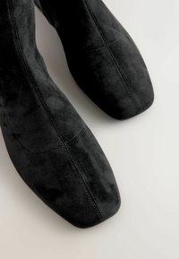 Sorte ruskinds ankler støvler med spids tå, der har en glat overflade og subtile stitch-detaljer langs siderne.