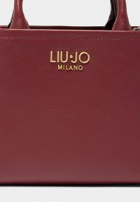 Sac à main en cuir bordeaux avec finition texturée, poignées doubles et accent logo doré "LIU•JO MILANO" sur le devant.
