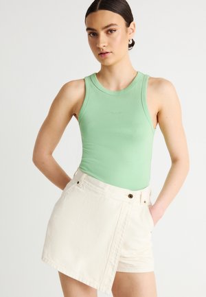 Ung kvinde med mørkt hår sat op i en knold, iført en ærmeløs, lysegrøn top og asymmetriske hvide shorts med hænderne i lommerne.