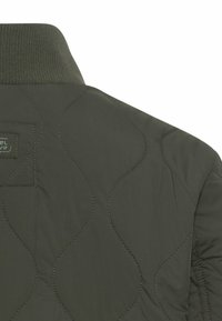 Tył oliwkowozielonej pikowanej kurtki z przeszyciami w kształcie fal oraz prążkowany kołnierz z małą czarną metką z tkaniny.