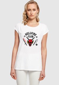 Camiseta de algodón blanca con un diseño gráfico de un diablo rojo con cuernos, el texto "HELLFIRE CLUB" y motivos decorativos de espada y látigo.