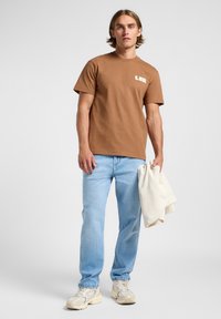 Camiseta de algodón marrón con mangas cortas y un logo; combinada con jeans rectos azul claro y zapatillas beige, sosteniendo una toalla crema.