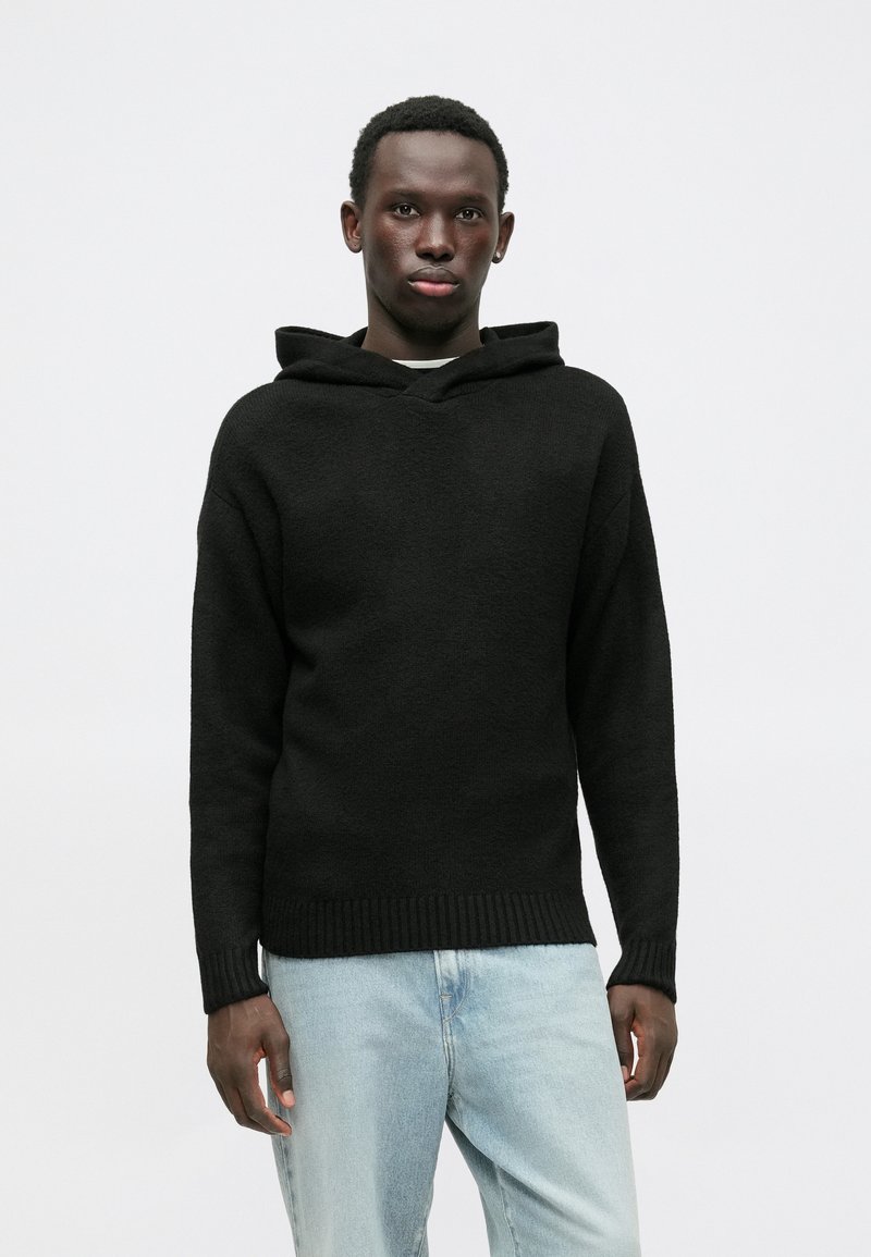 Black hooded sweater with a textured knit, featuring a loose fit and ribbed hem, paired with light blue jeans. Simple and functional design.