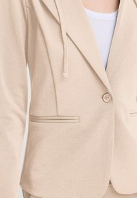 Béžový blazer s textúrovanou tkaninou, klopovými goliermi, zapínaním na gombík a jemným prúžkovaným akcentom na vrecku. Obsahuje kapucňu so sťahovacou šnúrkou.