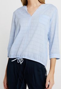 Helles blaues, strukturiertes Oberteil mit V-Ausschnitt, Dreiviertelärmeln und einer Brusttasche. Verfügt über einen Kordelzugsaum für eine verstellbare Passform.
