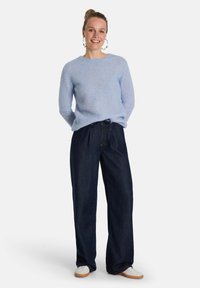 Hellblau gestrickter Pullover mit gerippter Textur, kombiniert mit dunkelblauen weit geschnittenen Jeans und weißen Sneakers. Der Pullover bietet eine lockere Passform und hat einen Kordelzug in der Taille.