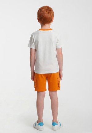 Dziecko z rudymi włosami stojące tyłem, ubrane w białą koszulkę, pomarańczowe szorty z kieszeniami oraz białe buty sportowe z niebieskimi akcentami.