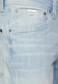 Los jeans de mezclilla de color azul claro presentan un corte clásico, un ligero desvanecimiento y costuras en contraste. Incluyen un bolsillo trasero con remaches de metal y una etiqueta de la marca.