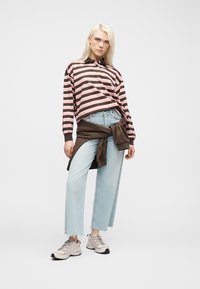 Νεαρή γυναίκα που φοράει ροζ και καφέ ριγέ μπλούζα με μακριά μανίκια, ανοιχτό μπλε παντελόνι με φαρδιά πόδια, μπεζ αθλητικά παπούτσια και καφέ φούτερ δεμένο στη μέση.