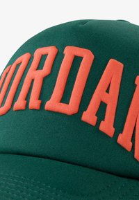 Gorra verde con el prominente texto en naranja "JORDAN". El material es una tela suave; el diseño presenta una forma estructurada y acentos cosidos.