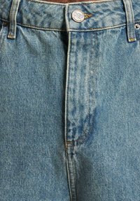 Jeans de mezclilla azul claro con una textura desgastada, cuentan con un botón plateado con "DEF" grabado y costuras anaranjadas a lo largo de las costuras.
