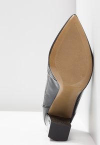 Botte en cuir noir à bout pointu avec une semelle texturée beige et un talon bloc, vue par le dessous.