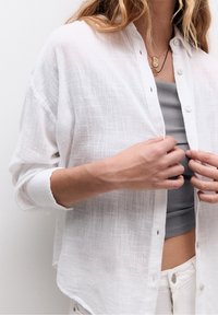 Witte, doorzichtige button-up blouse van een gestructureerde stof. Heeft een comfortabele pasvorm, verlaagde schouders en opgerolde mouwen. Gedragen over een grijze top.