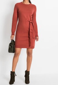 Abito lavorato a maglia color ruggine con maniche lunghe, dettaglio alla vita con nastro e orlo a coste. Indossato con una borsa a tracolla verde oliva e stivaletti neri.