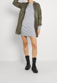 Olivgrön glänsande konstläder skjortjacka över en ljusgrå stickad klänning; ihopparat med knähöga svarta snörstövlar.