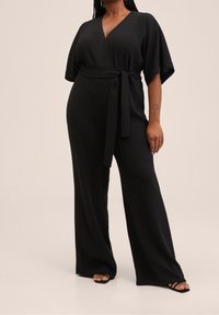 Combinaison noire avec un décolleté en V, des manches larges et courtes, et une ceinture à nouer à la taille. Le tissu semble doux et texturé, avec un pantalon à jambes larges.