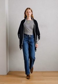 Schwarzer Fransen-Schal über einem schwarz-weiß gestreiften Oberteil, kombiniert mit hoch taillierten blauen Jeans und braunen Wildleder-Stiefeletten.