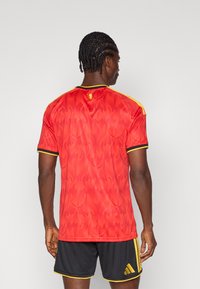 Man met gevlochten haar staat met zijn rug naar de camera, gekleed in een rood en geel sportshirt met vlammenpatronen en zwarte korte broek met gele strepen.