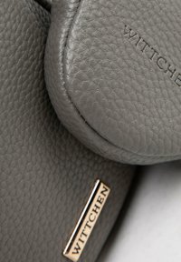 Graune strukturierte Lederhandtasche mit geprägten und metallenen "Wittchen"-Markenetiketten, die auf überlappenden Bereichen sichtbar sind.
