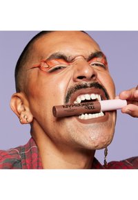 NYX lingerie XXL läppprodukt hålls i tänderna, brun tub med rosa kork. Ämnet har kort hår, ansiktsbehåring, orange eyeliner och rutig skjorta.