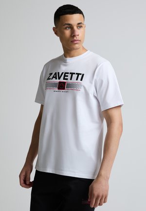 Ung mand med kort mørkt hår iført en hvid Zavetti T-shirt med sort og rødt grafisk motiv og sorte shorts på en ensfarvet grå baggrund.