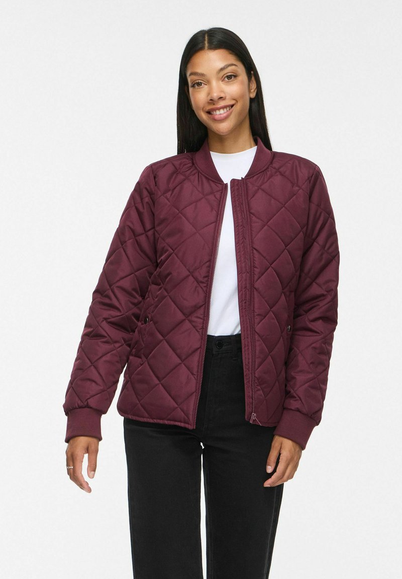 Femme souriante portant une veste matelassée bordeaux par-dessus une chemise blanche et un pantalon noir sur un fond clair uni.