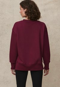 Burgundská mikina s vroubkovaným kulatým výstřihem, dlouhými rukávy a uvolněným střihem. Obsahuje mírně zaoblený lem a měkkou texturu.