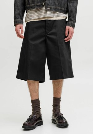 Mann iført svarte, vide knelange shorts, brune sokker, mørkebrune tasselloafers, rutete skjorte og mørk denimjakke.
