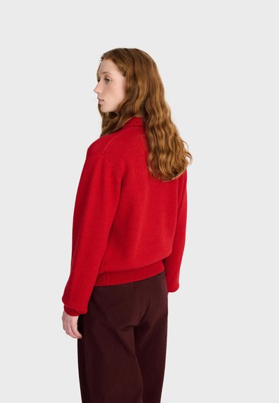 Jeune femme aux longs cheveux roux ondulés, portant un pull rouge et un pantalon foncé, debout de profil devant un fond clair uni.