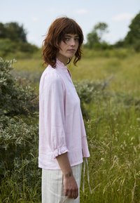 Jeune personne aux cheveux roux portant une blouse rose clair et un pantalon blanc, debout dans un champ d'herbe verte, regardant vers la caméra.