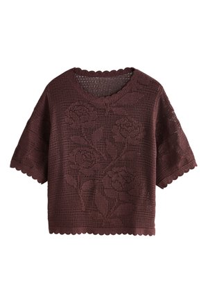 Mladá žena s tmavými kučeravými vlasmi má na sebe tmavý pletený top a dlhú sukňu s čipkovaným lemom, opiera sa o béžovú textúrovanú stenu pri zdobenej okennej mreži.