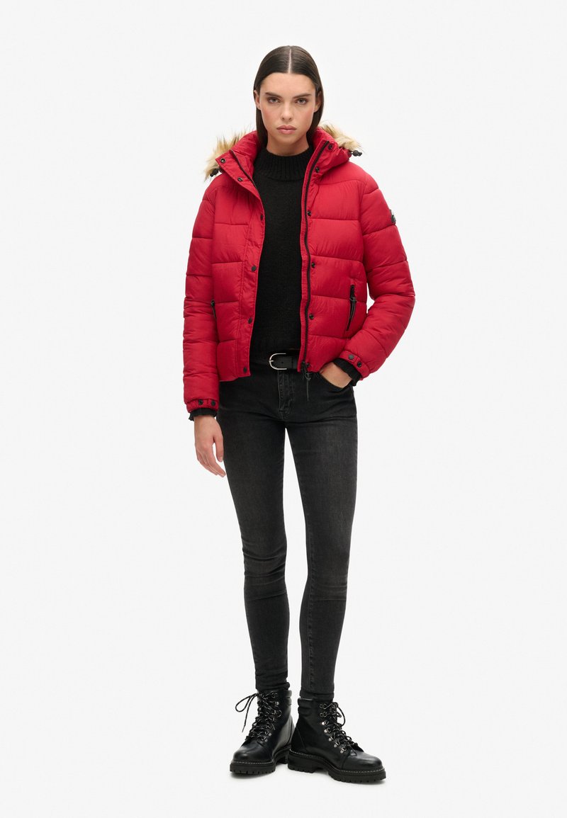 Giacca puffer rossa con cappuccio foderato in pelliccia rimovibile, apertura con zip e due tasche laterali, abbinata a jeans skinny neri e stivali con lacci.